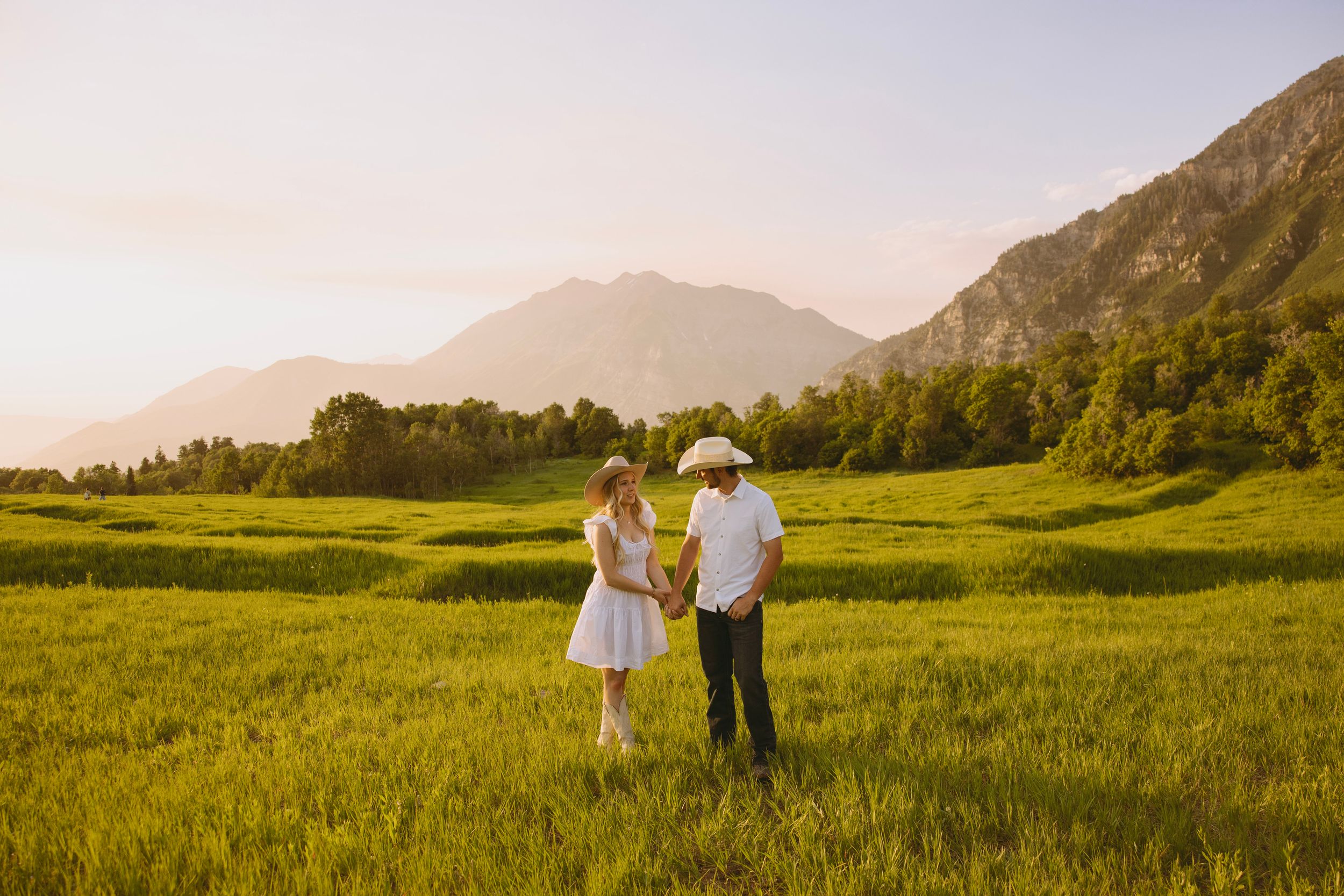 Couple in white clothing walking through a scenic mountain meadow during golden hour.