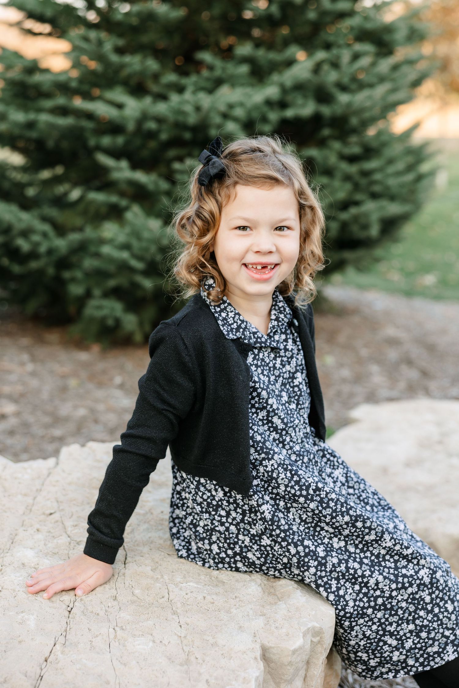 A small child sits on a rock wearing a black cardigan over a floral print dress near an evergreen tree.
