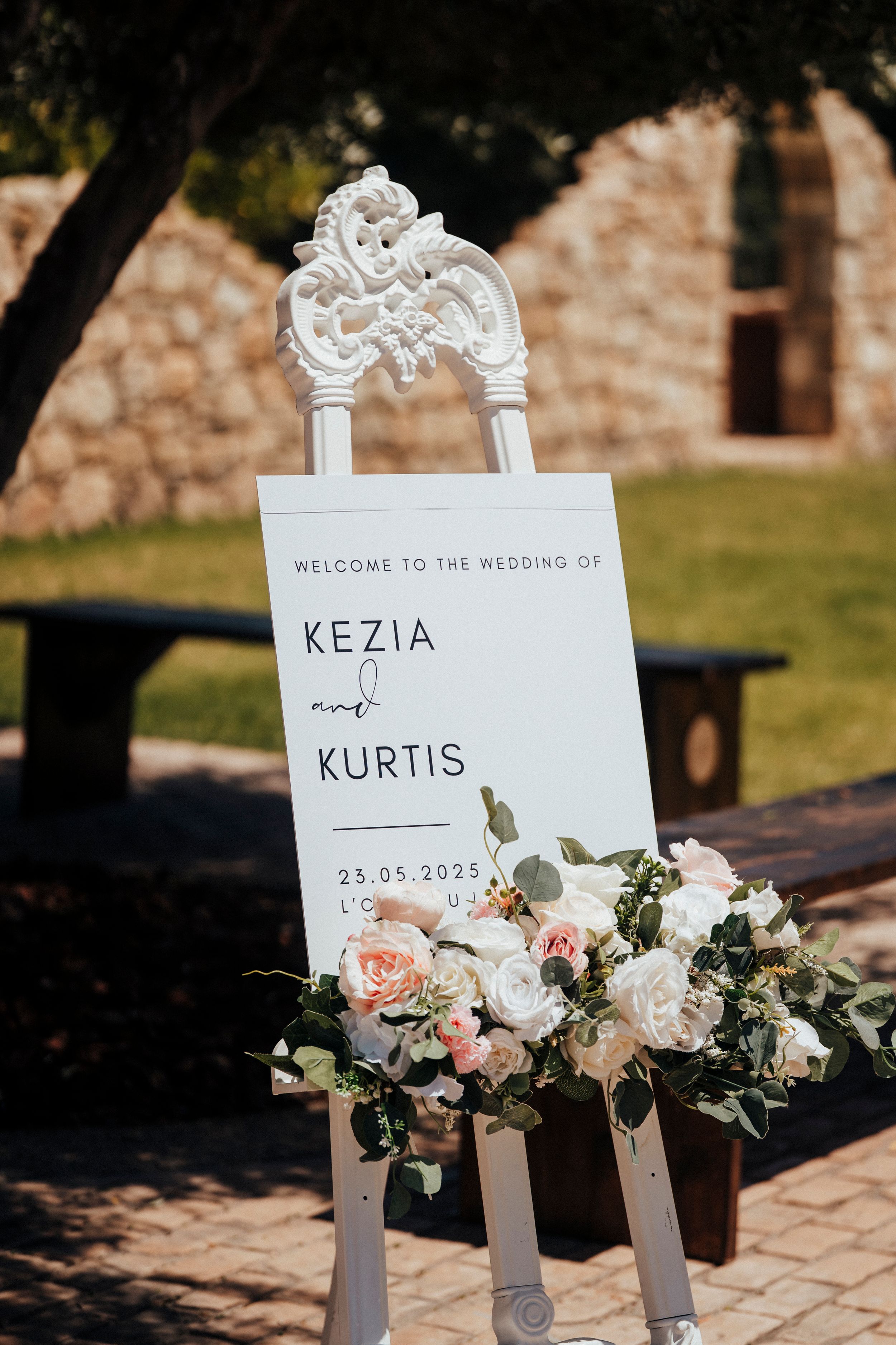 Wedding welcome sign with ornate white frame displays Kezia and Kurtis's names with blush and white floral arrangement below.