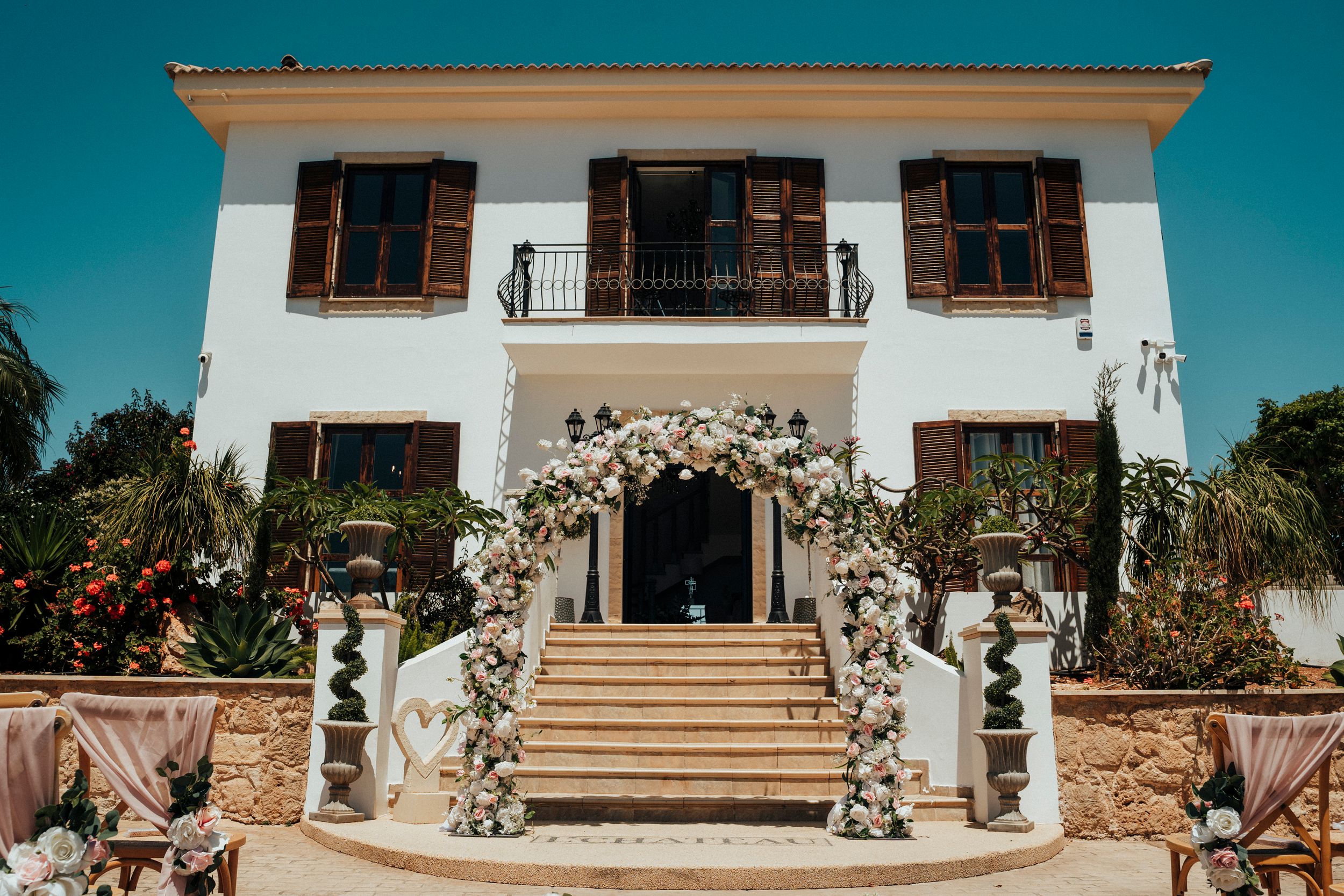 A wide panoramic view of a Mediterranean-style white villa with flowering gardens and wooden balconies against a blue sky.