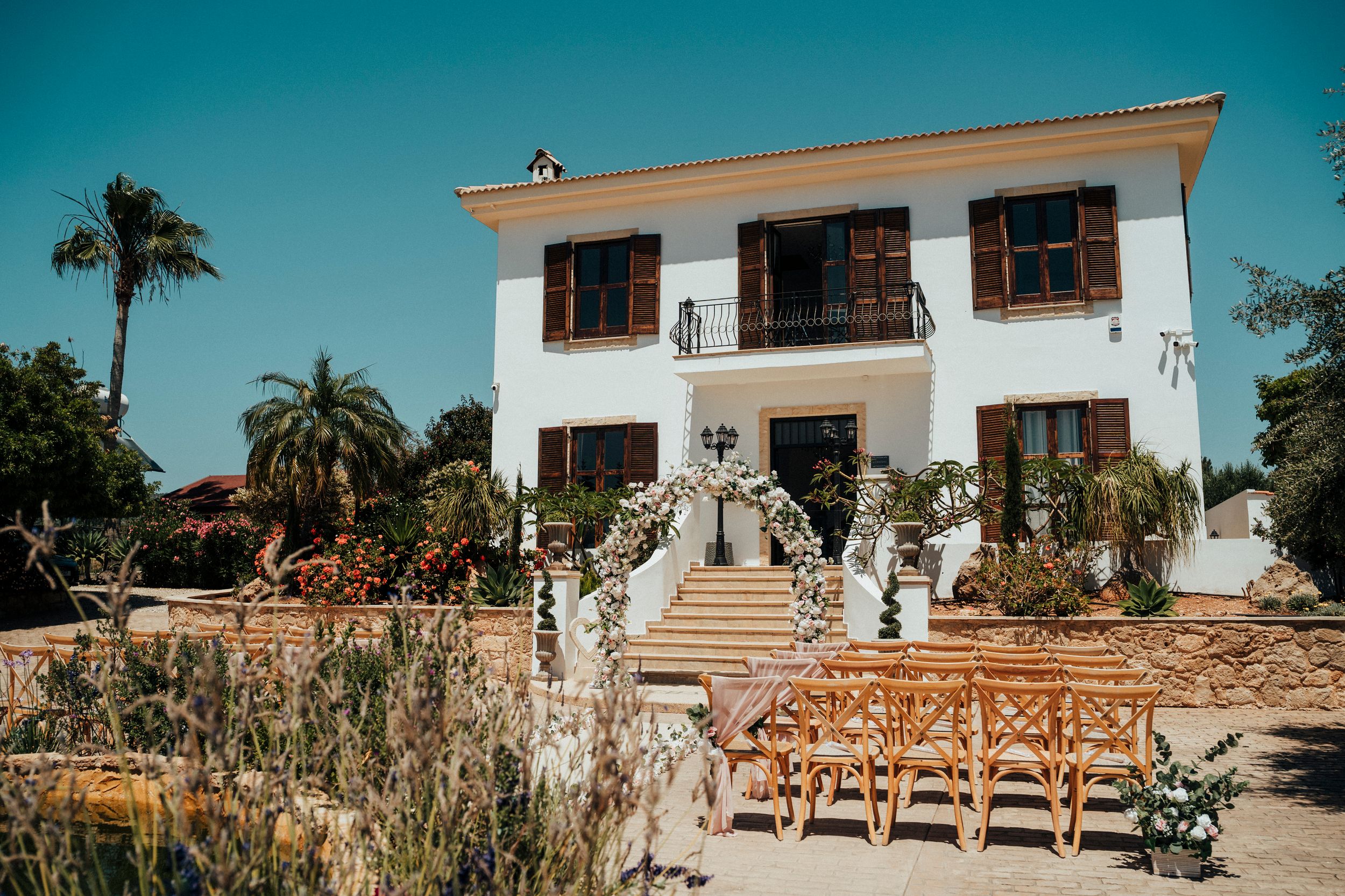 Panoramic view of Spanish-style outdoor wedding venue with palm trees and wooden chairs facing a white villa.