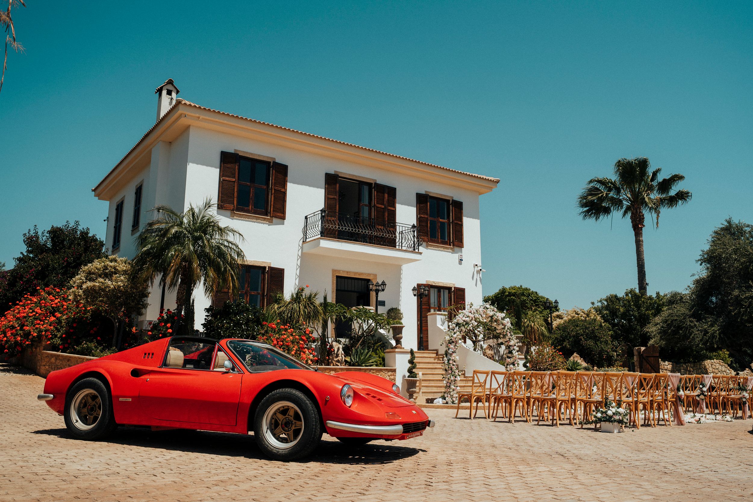 Five classic vintage cars lined up outside a Mediterranean-style villa with palm trees against a clear blue sky.