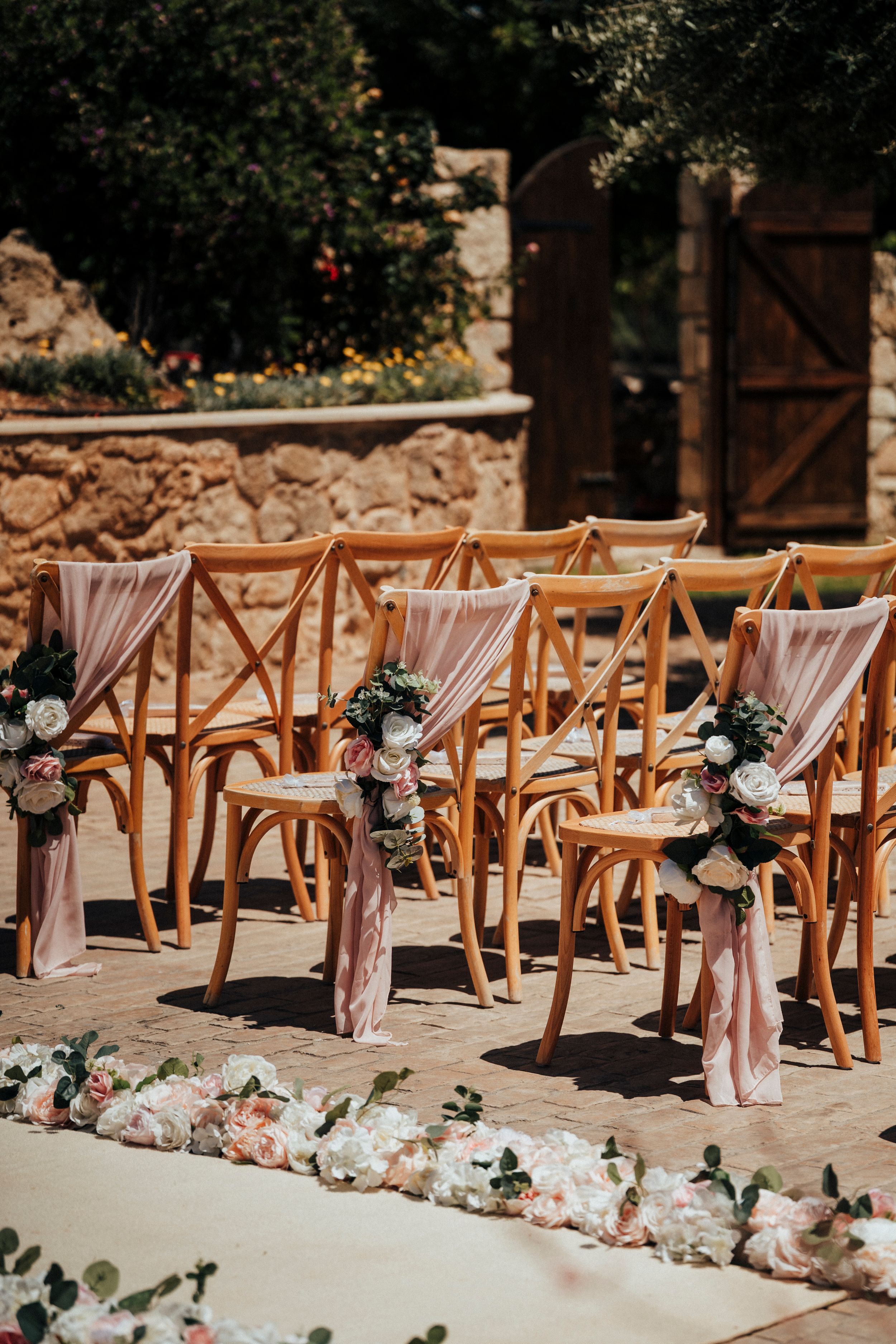 Rows of wooden chairs with pink fabric draping and floral arrangements line a sunny outdoor ceremony space at a wedding.