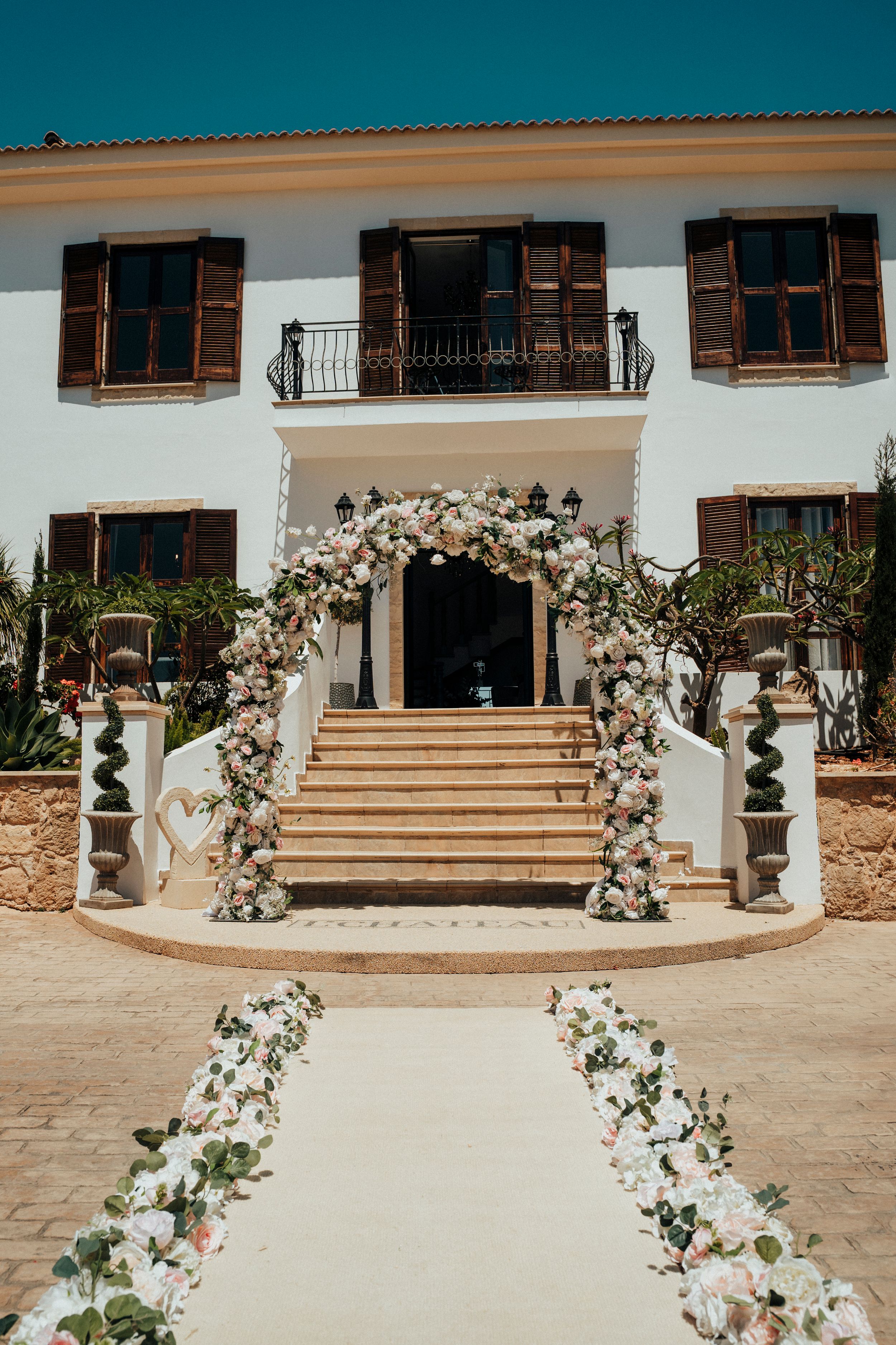 Wedding ceremony setup with curved floral arches creating a path to elegant stone steps at a Mediterranean-style venue.