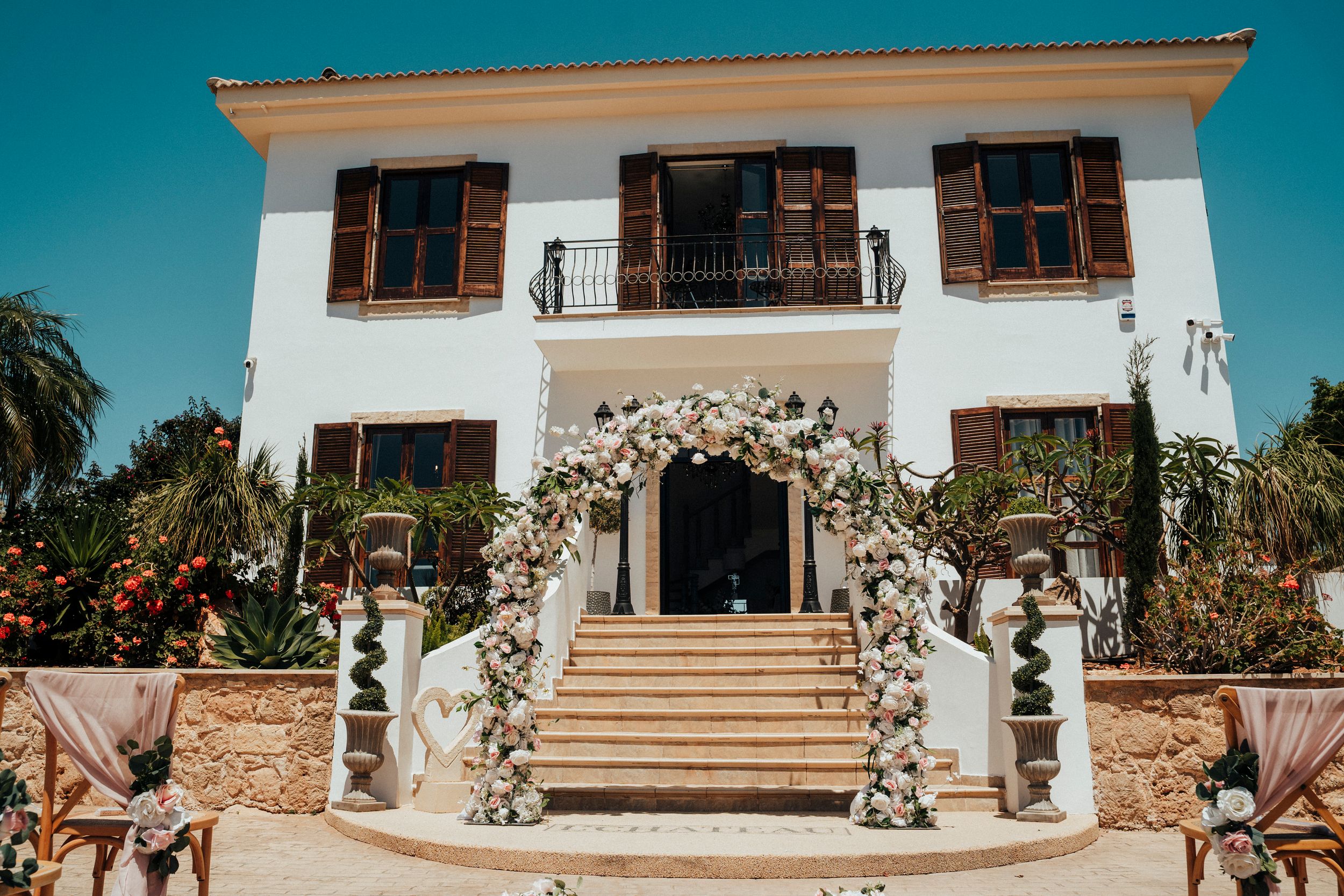 Elegant white two-story mansion with wooden shutters and grand floral archway entrance decorated for a wedding ceremony.