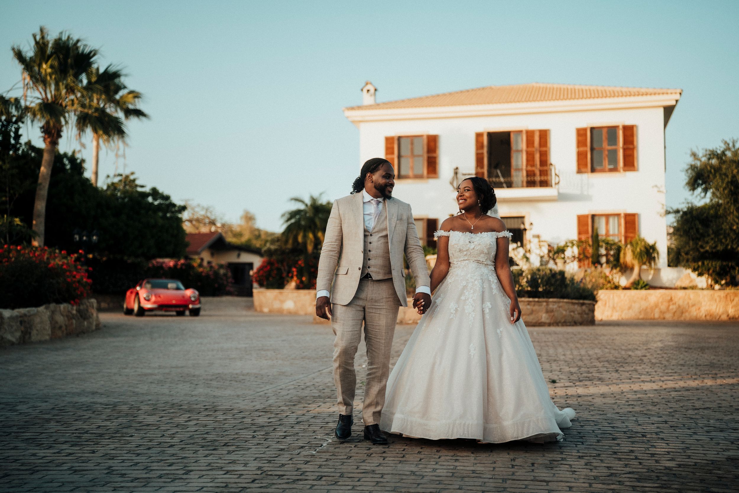 Newlywed couple walks hand in hand on a cobblestone driveway in front of Mediterranean villa at sunset.