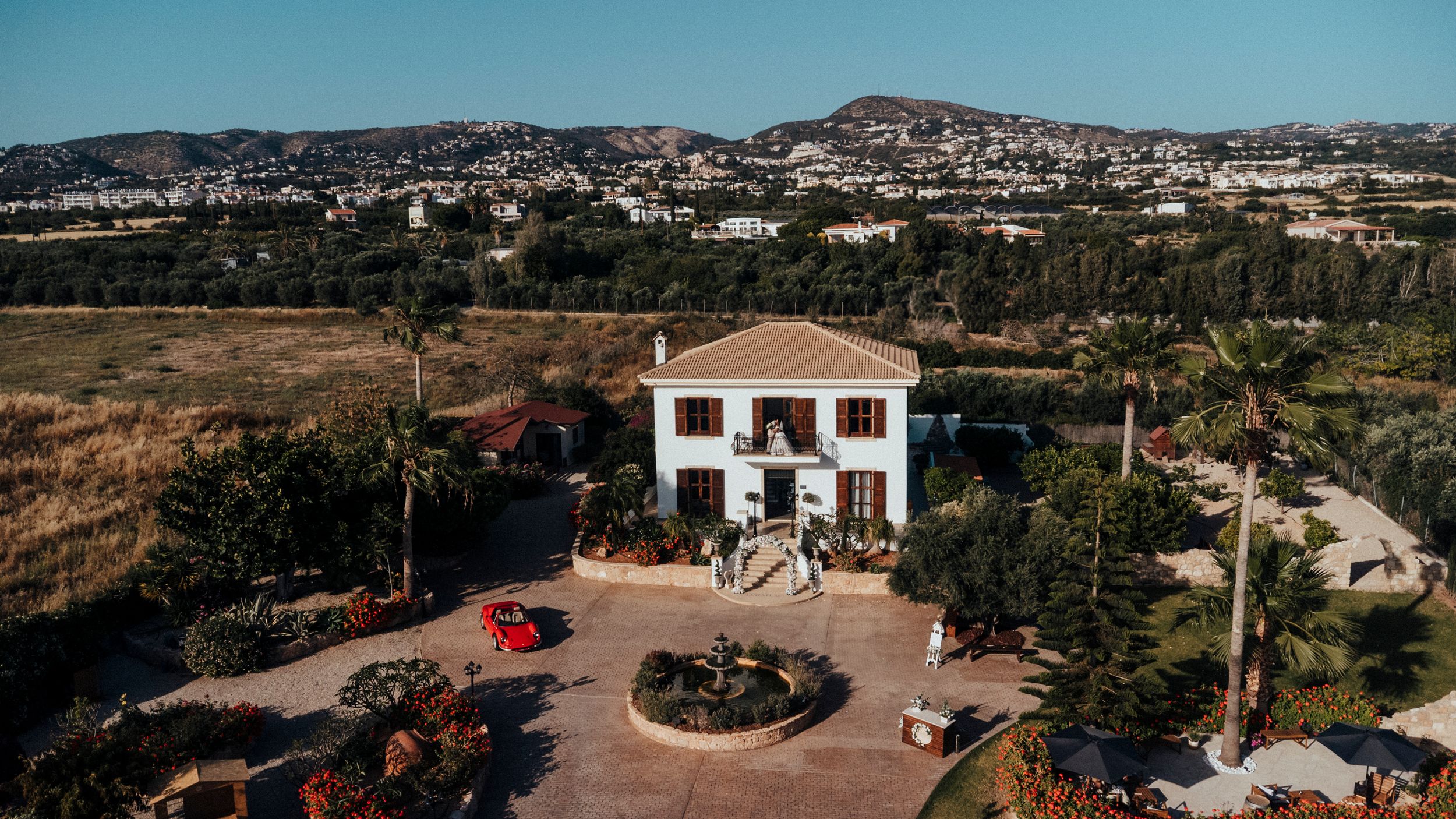 Aerial view of a white Mediterranean villa with red car in driveway surrounded by arid landscape and distant hills.