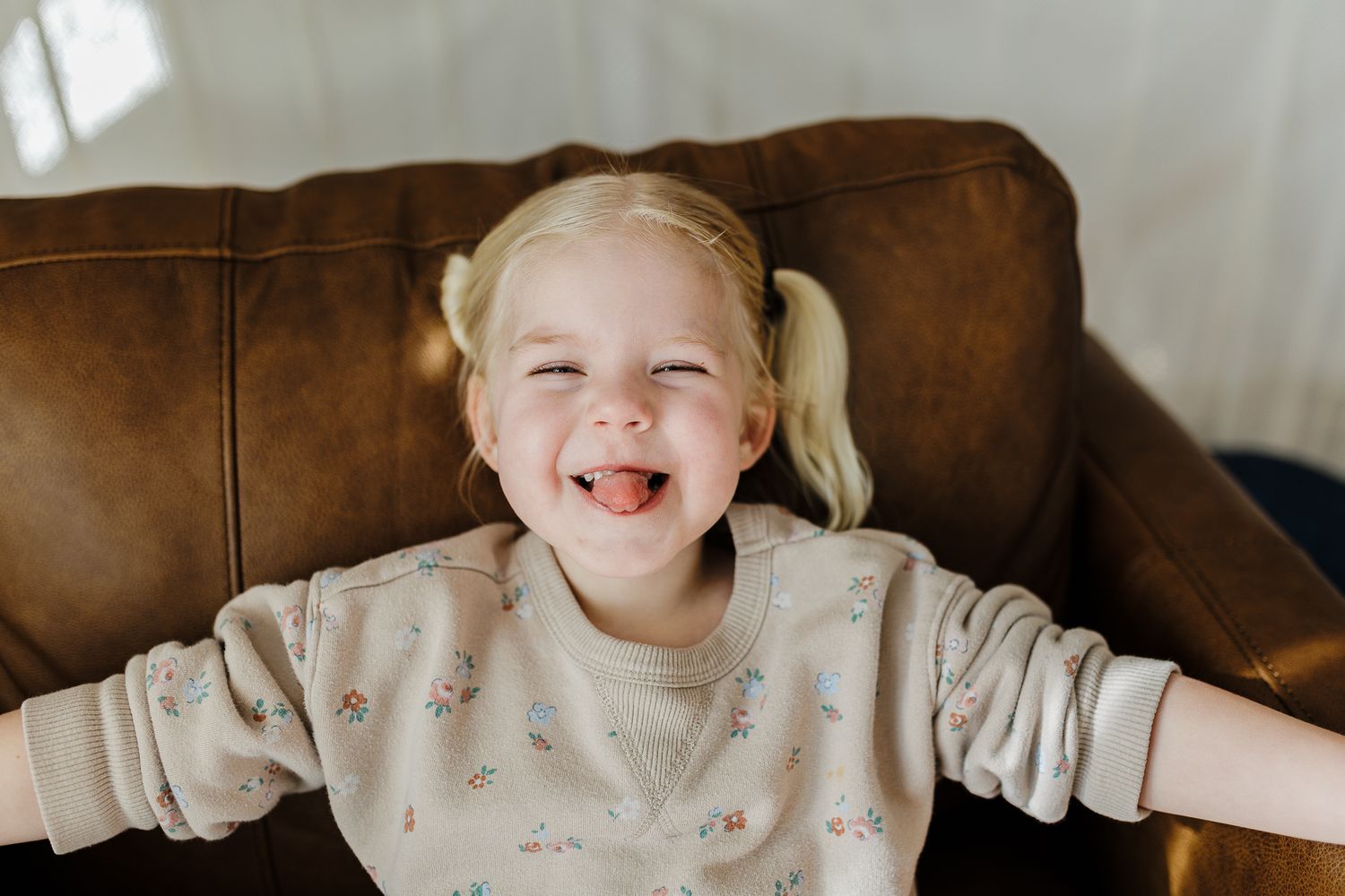 Young child with blonde pigtails wearing a patterned beige sweatshirt laughs joyfully while sitting on brown leather couch.