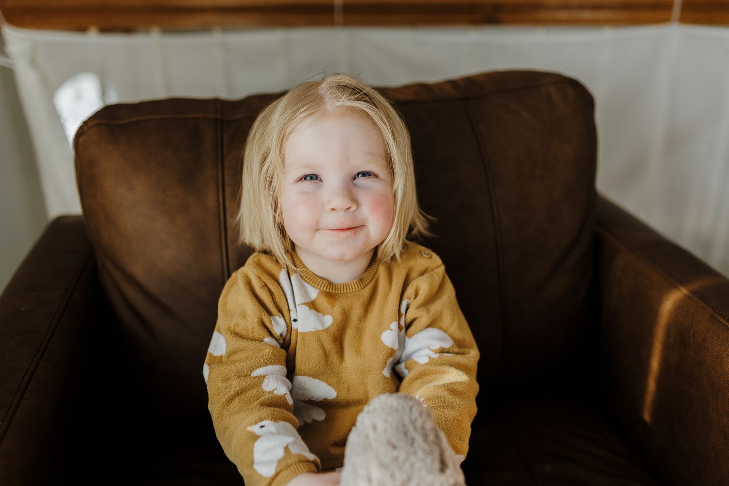 Child in mustard yellow sweatshirt with stars sits contentedly on brown leather sofa against white wall.