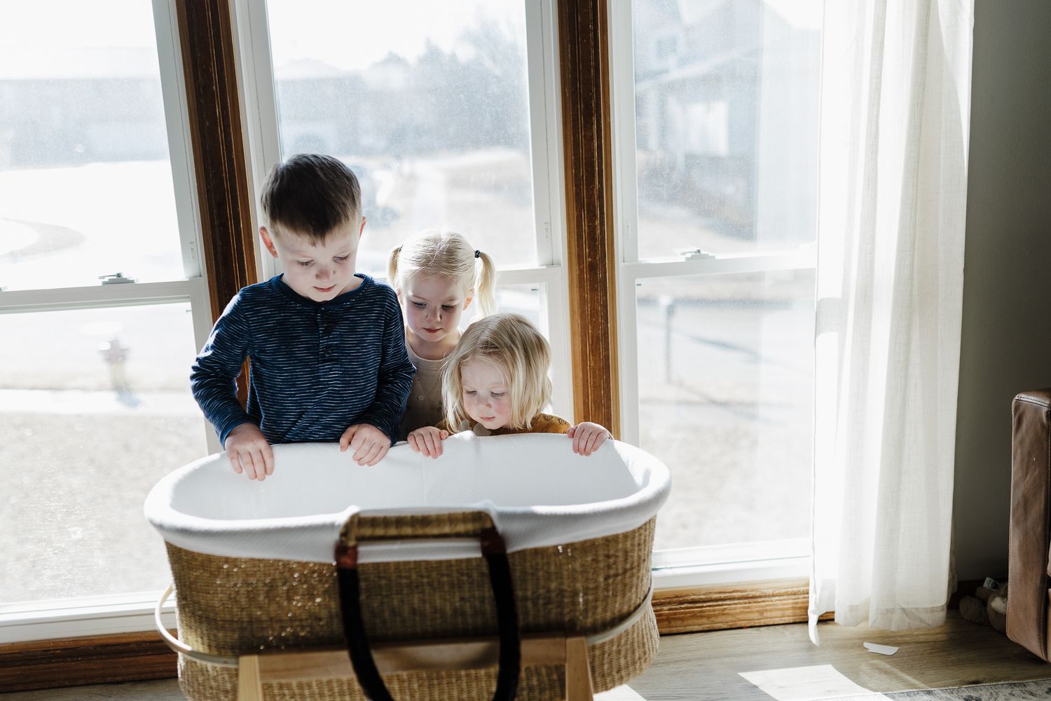 Three children share a cozy reading moment together in an oversized cushioned chair by the window.