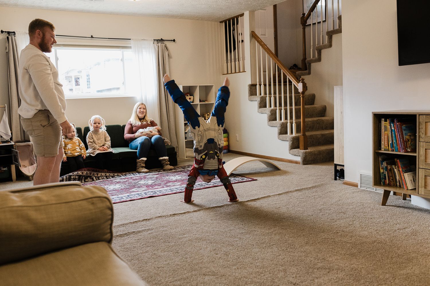 A child does a handstand in a cozy living room with carpet, stairs, and bookshelves while others watch from the sofa.