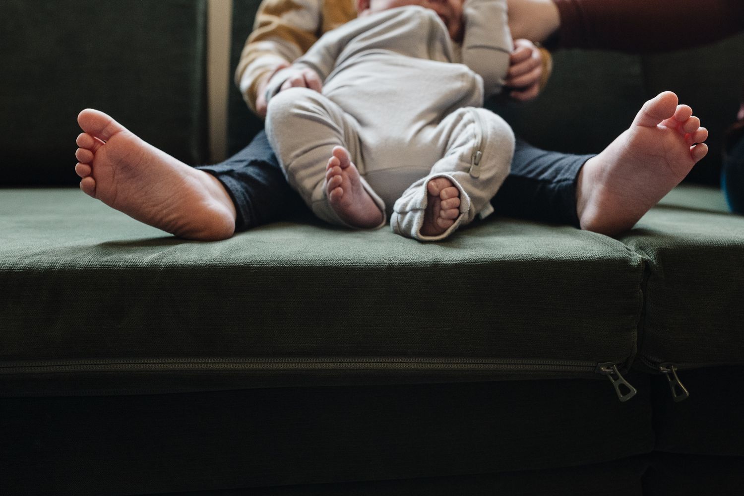 Close-up view of tiny bare feet resting on a dark green couch cushion with white baby clothing visible.
