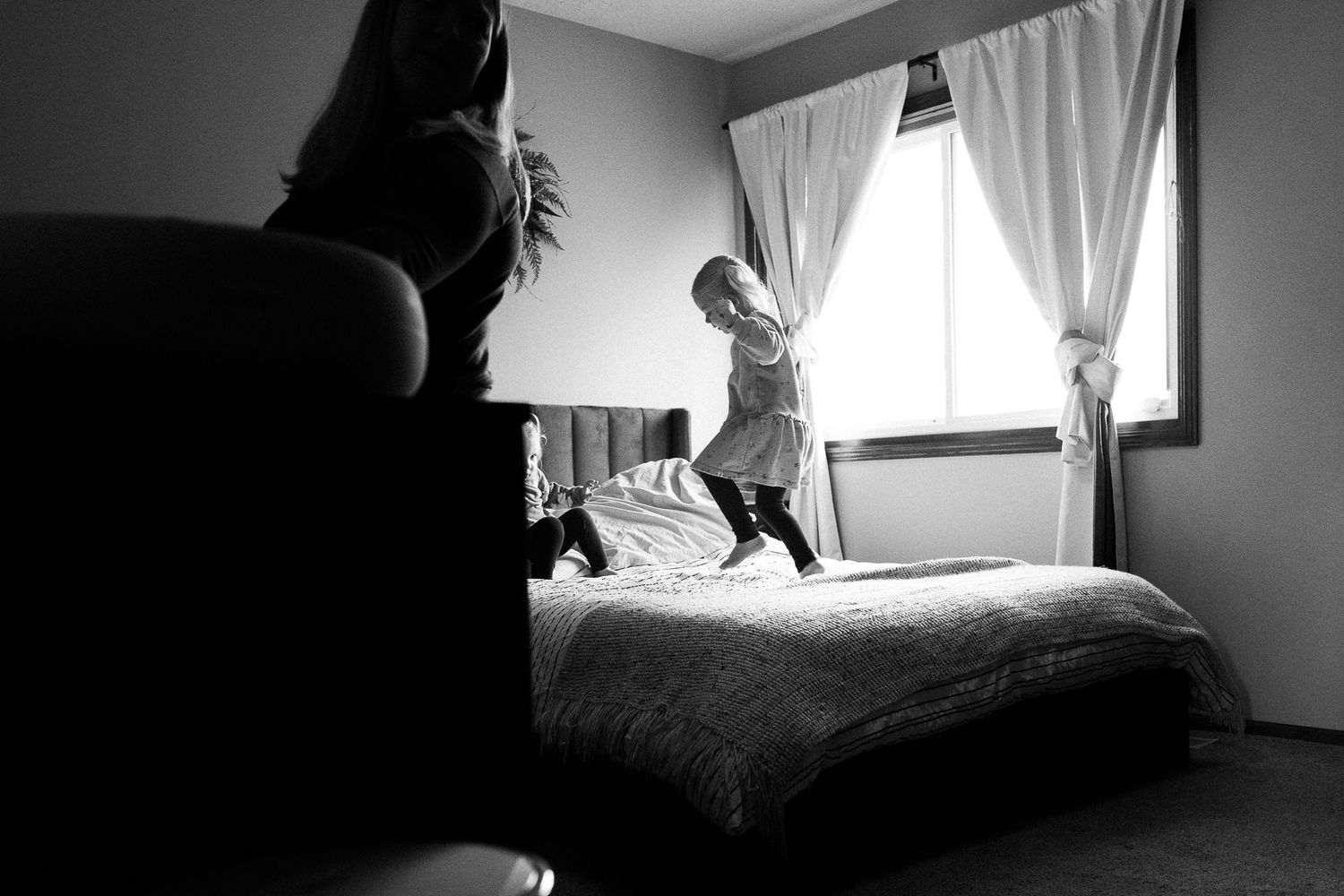 A silhouetted figure watches a small child jumping playfully on a bed near a curtained window in black and white.