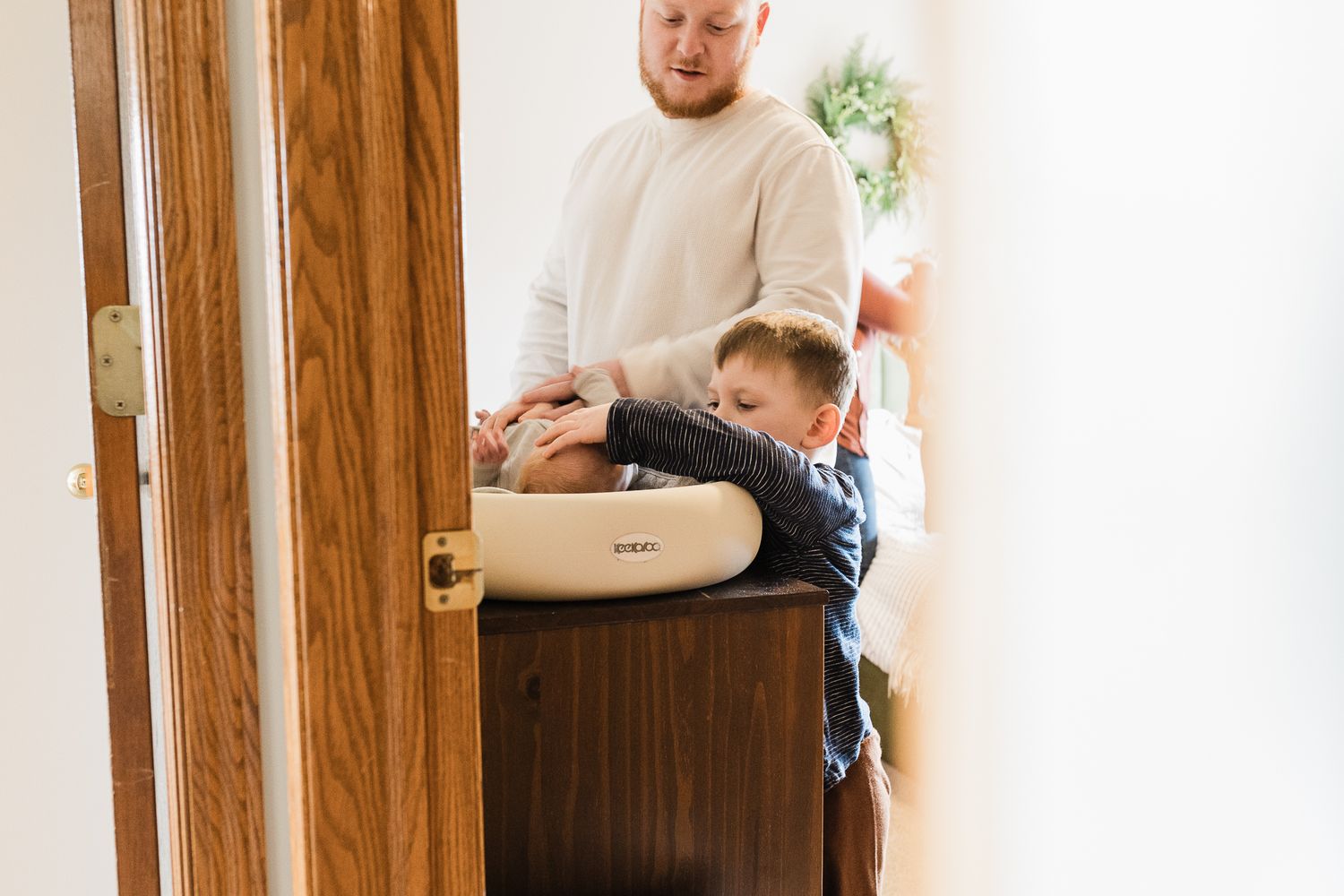 A child peeks through behind a wooden door while an adult in a cream sweater stands nearby.