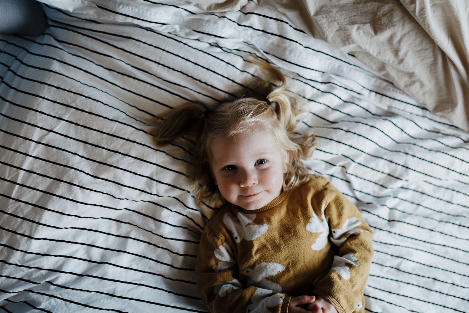Child lying on striped bedding wearing golden shirt with hair spread out on pillow.