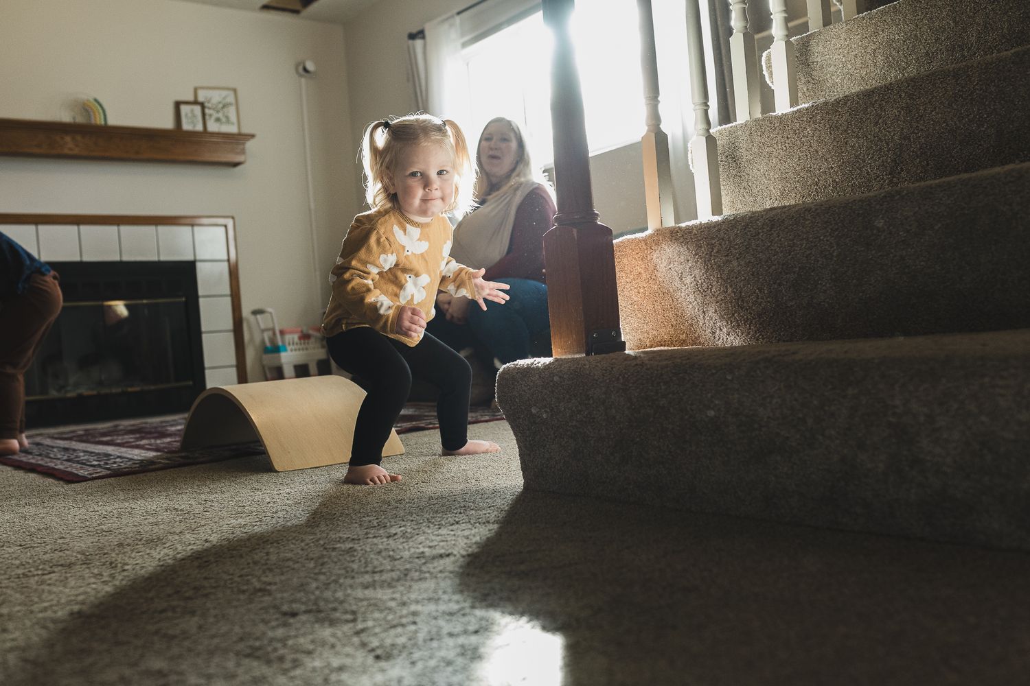 A toddler sits on carpeted stairs in a sunlit living room while holding a plush stuffed toy.