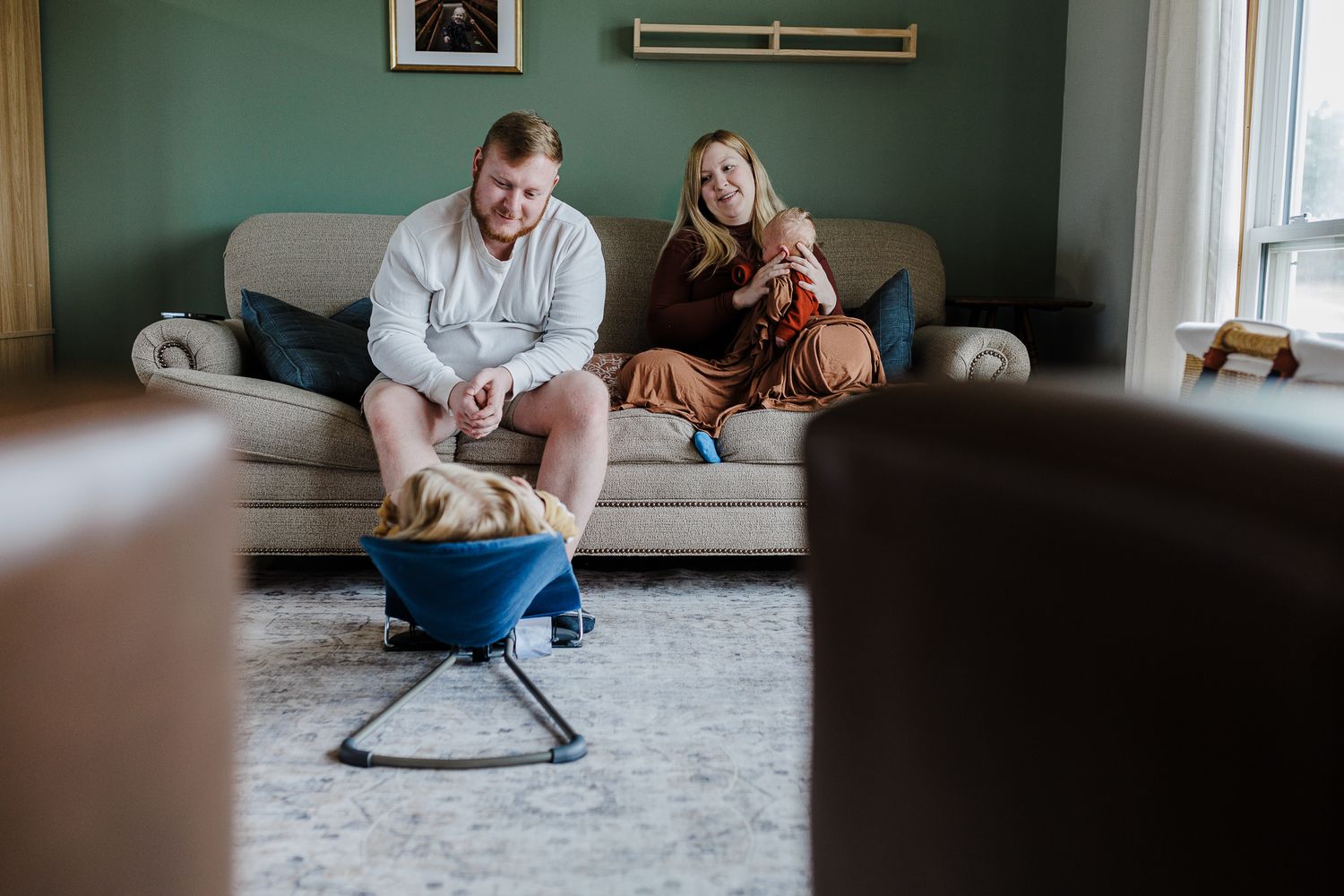 Two people relax on a grey couch while a baby rests in a blue bouncer seat near a mint green wall.