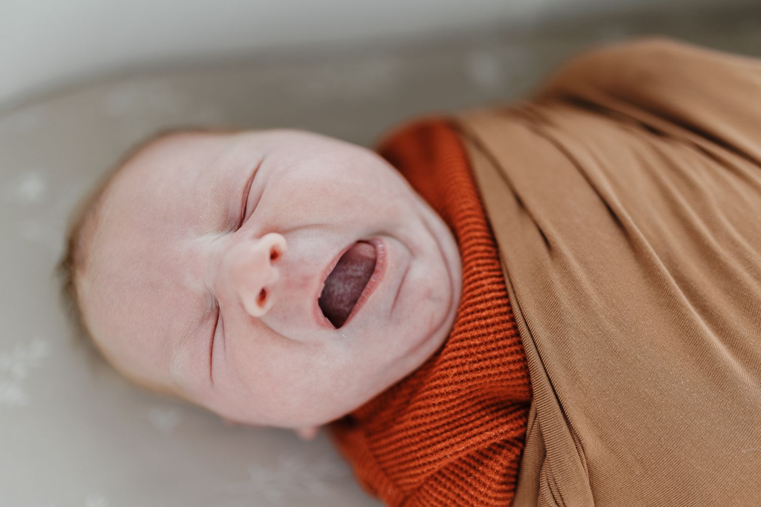 Close-up of a smiling baby wearing an orange and tan outfit laying on a light colored surface.