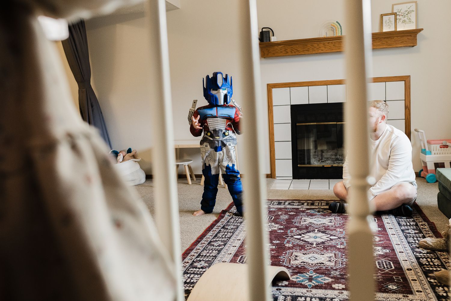 Small child wearing a metallic blue robot costume plays in a cozy living room with a fireplace and patterned area rug.
