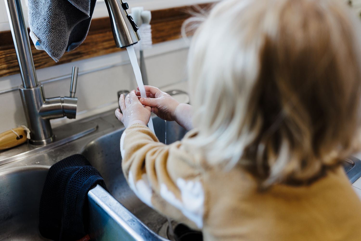 Child in beige sweater stands at kitchen sink learning to wash hands with flowing water from faucet.