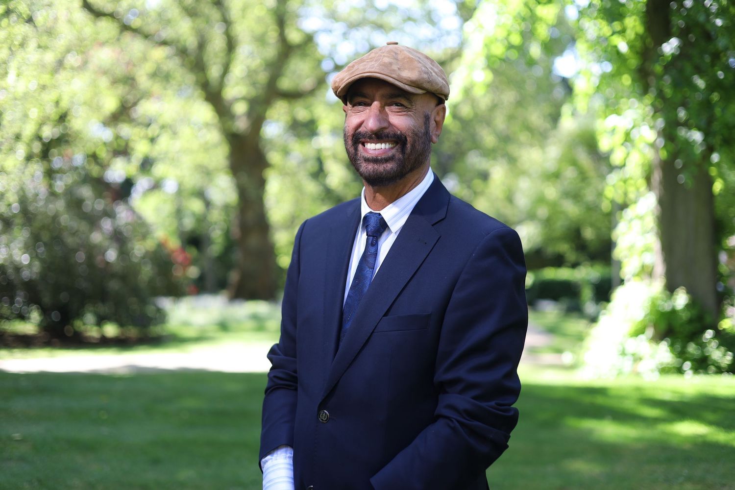 Professional in a navy suit and baseball cap smiles while standing in a sunny garden setting.