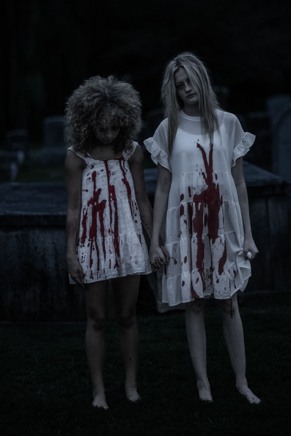 Two figures in white blood-stained dresses stand barefoot in a dark outdoor setting creating an eerie horror scene.