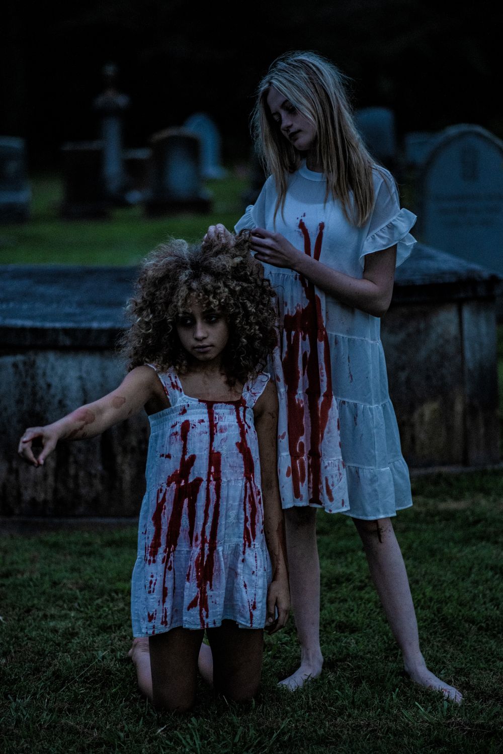 Two figures wearing white dresses with blood stains pose dramatically in a dark cemetery setting.