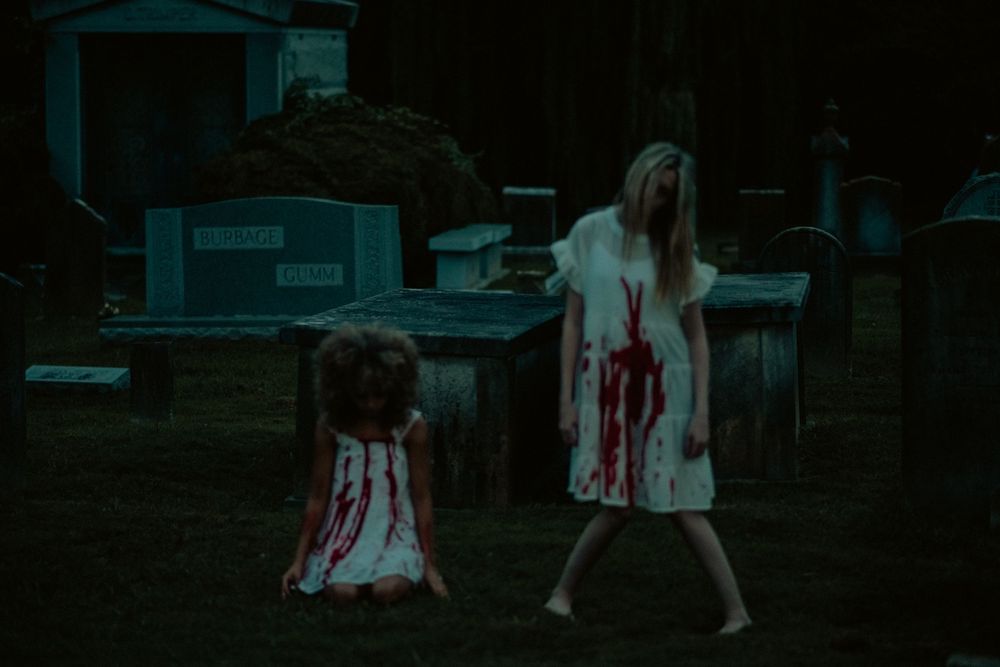 A figure in a blood-stained dress stands menacingly in a dark cemetery at night with gravestones visible around them.