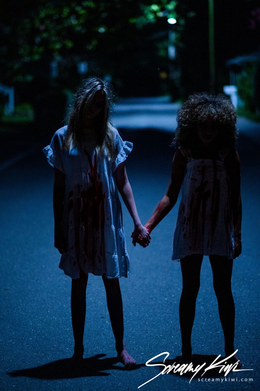Two figures in dresses hold hands while standing on a dark street at night creating a spooky scene.