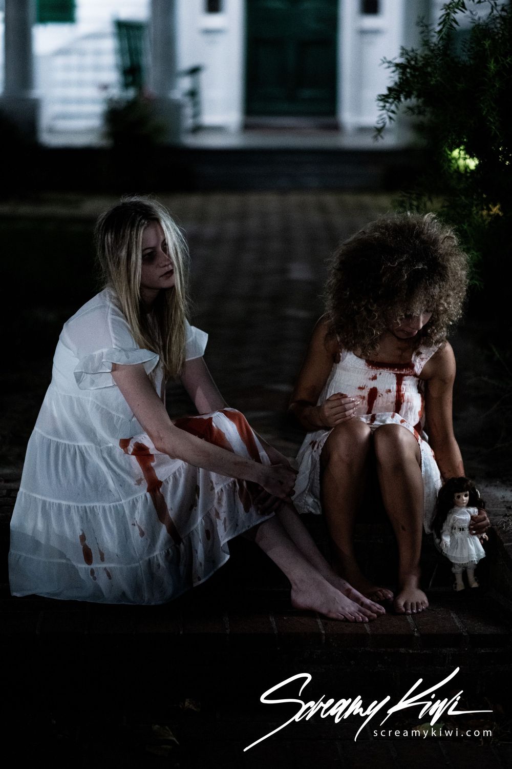 Two figures in white dresses sit together on steps at dusk with dramatic moody lighting.