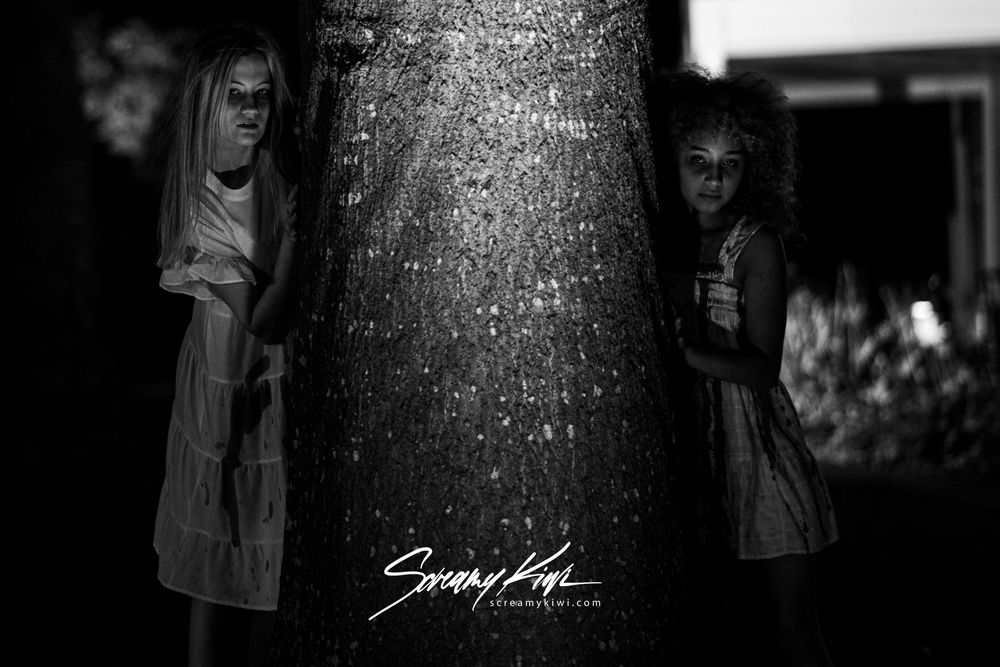 Two figures in white dresses peek out from behind a large tree trunk creating an unsettling black and white image.