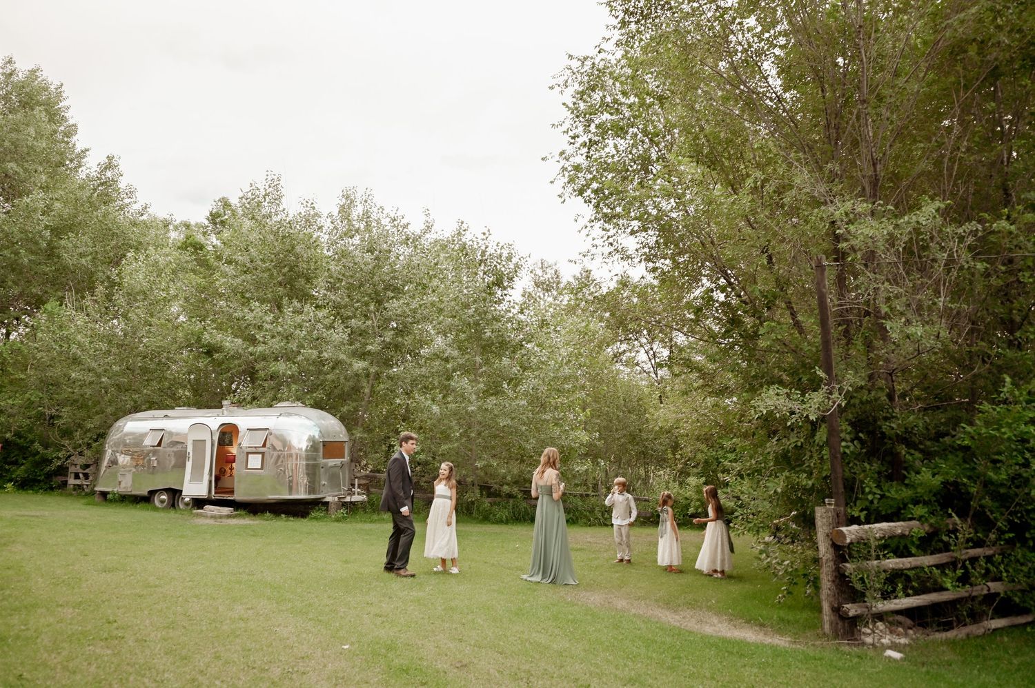 Vintage Airstream trailer parked on green lawn with wedding party gathered outside in pastoral woodsy setting.