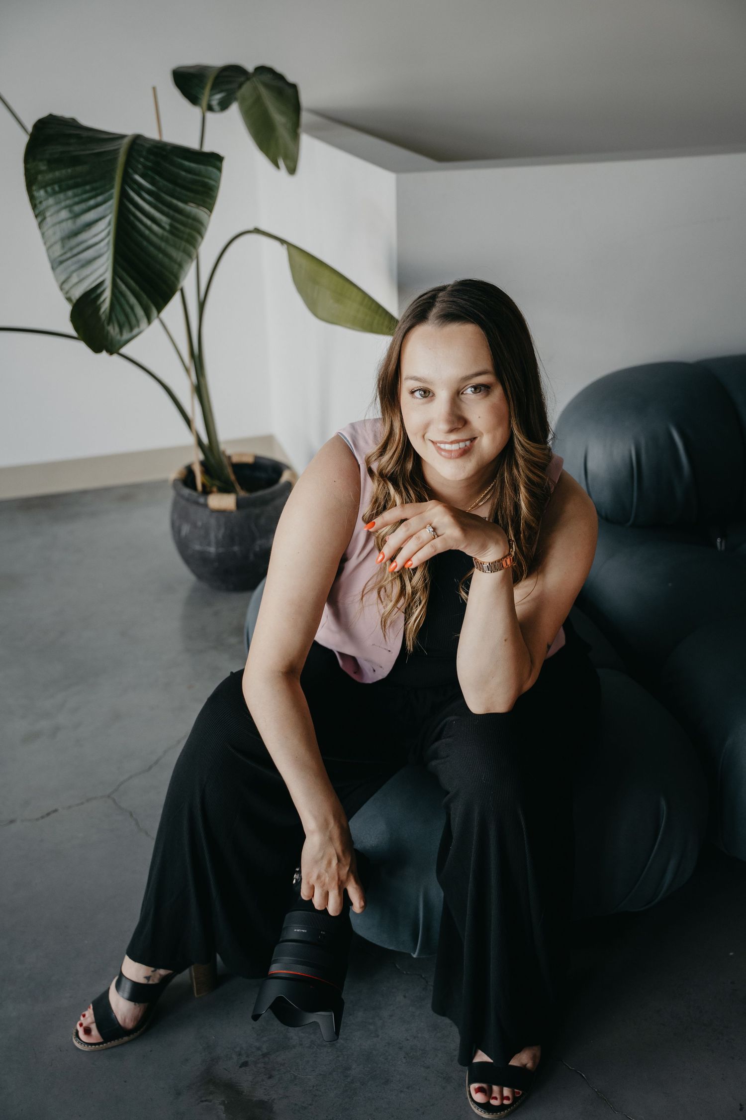 Person sitting in a black armchair wearing a pink blouse and black pants next to a large tropical houseplant in a modern space.