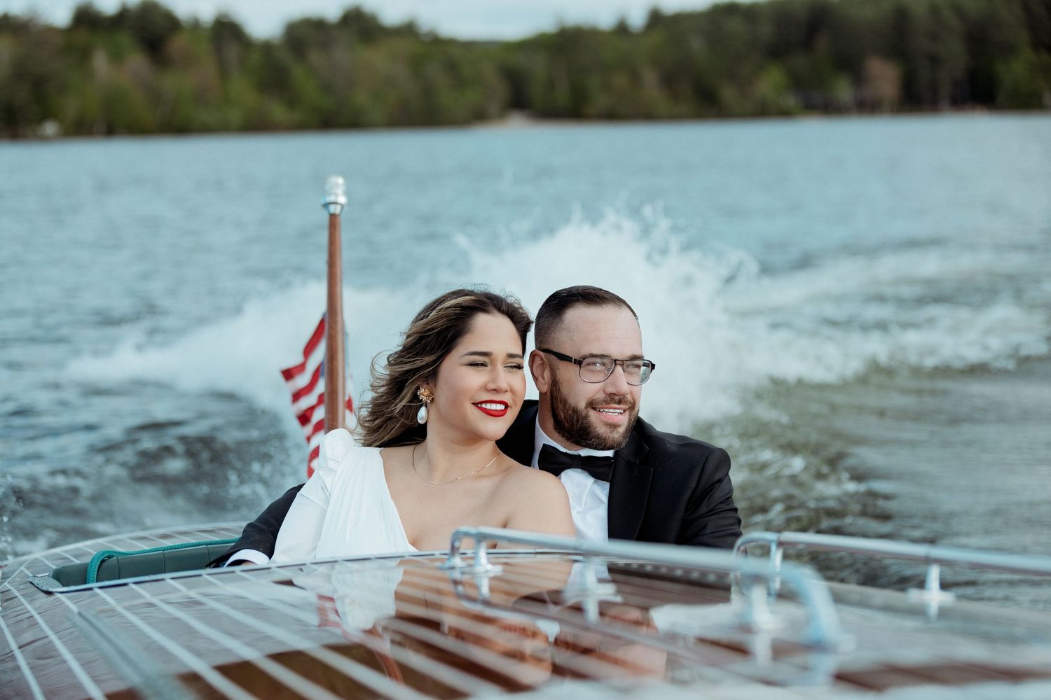 A couple smiles while riding in a vintage wooden boat on a lake with an American flag waving in the breeze.