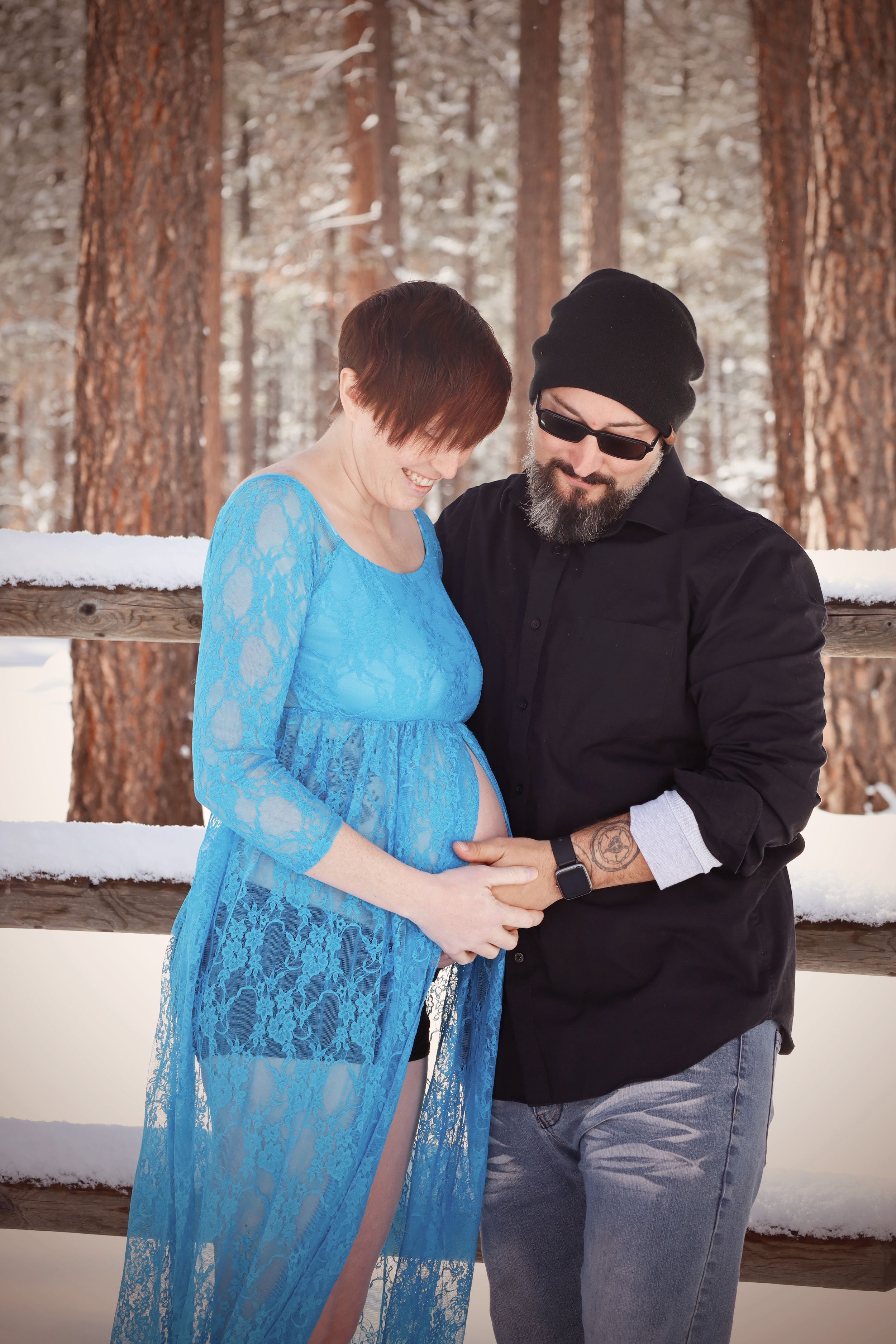 Pregnant woman in blue lace gown with bearded man in black holding her baby bump in snowy winter setting.