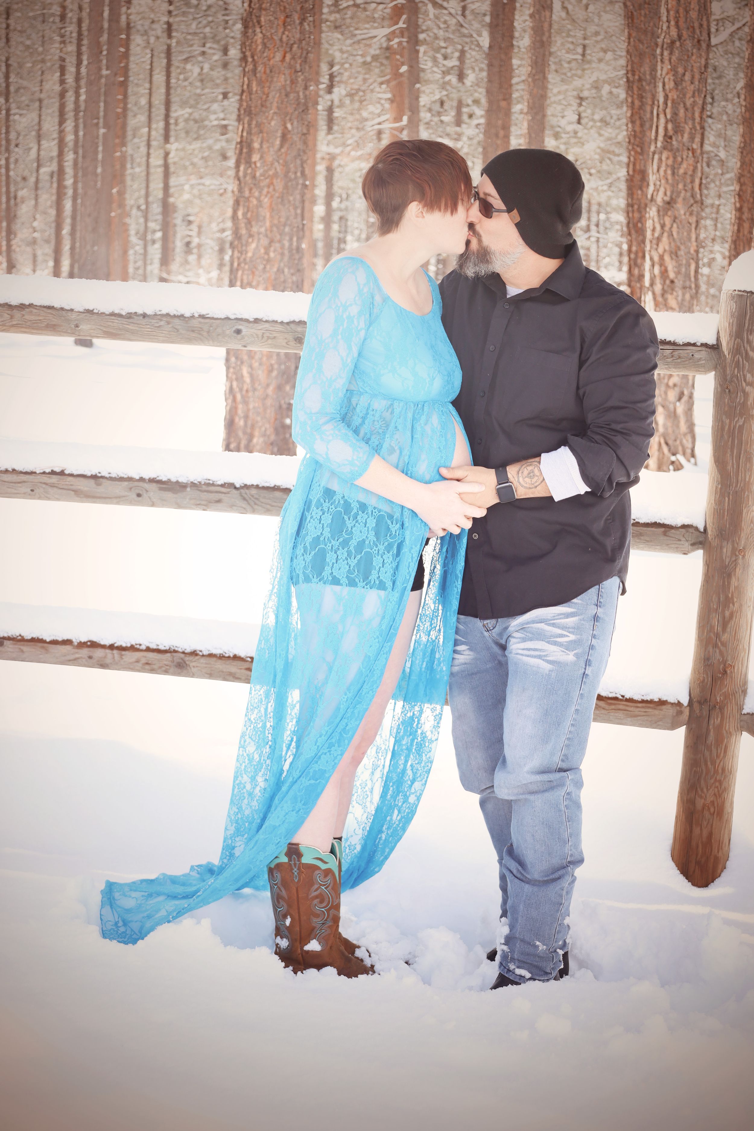 Couple sharing a tender kiss outdoors in snow, woman wearing blue lace maternity gown, man in black beanie.