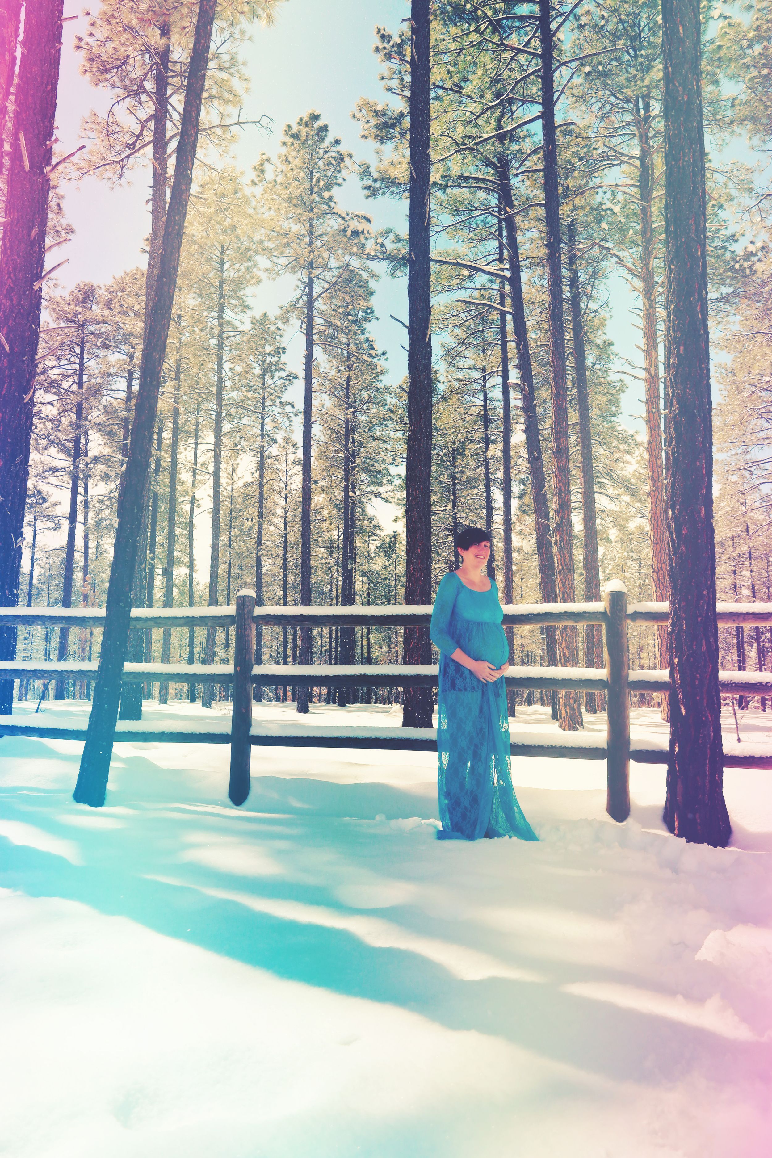 Pregnant woman in teal lace maternity gown standing in snowy pine forest with warm light.