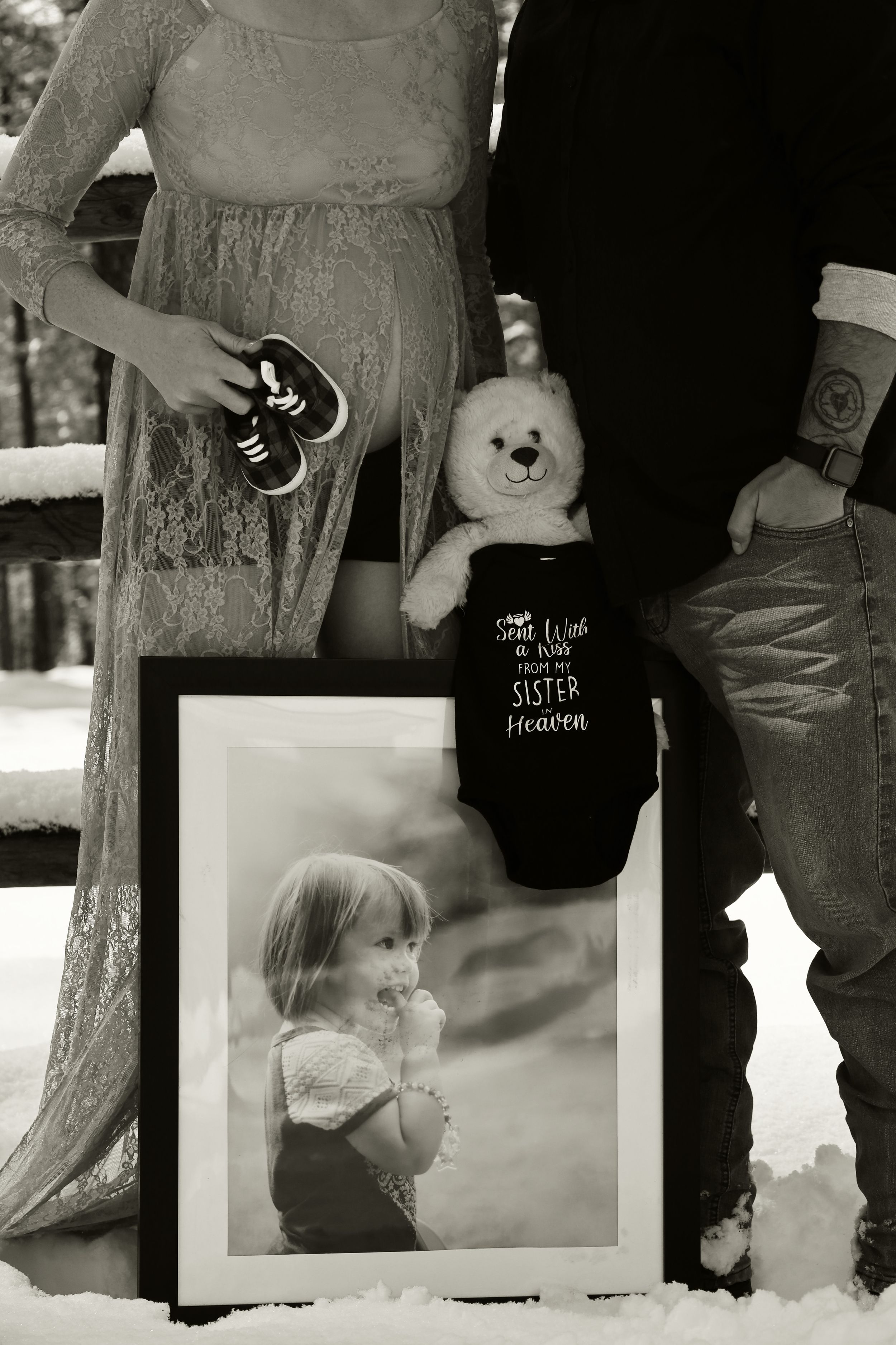 Black and white maternity photo with couple holding baby shoes, teddy bear, and framed child portrait in snow.