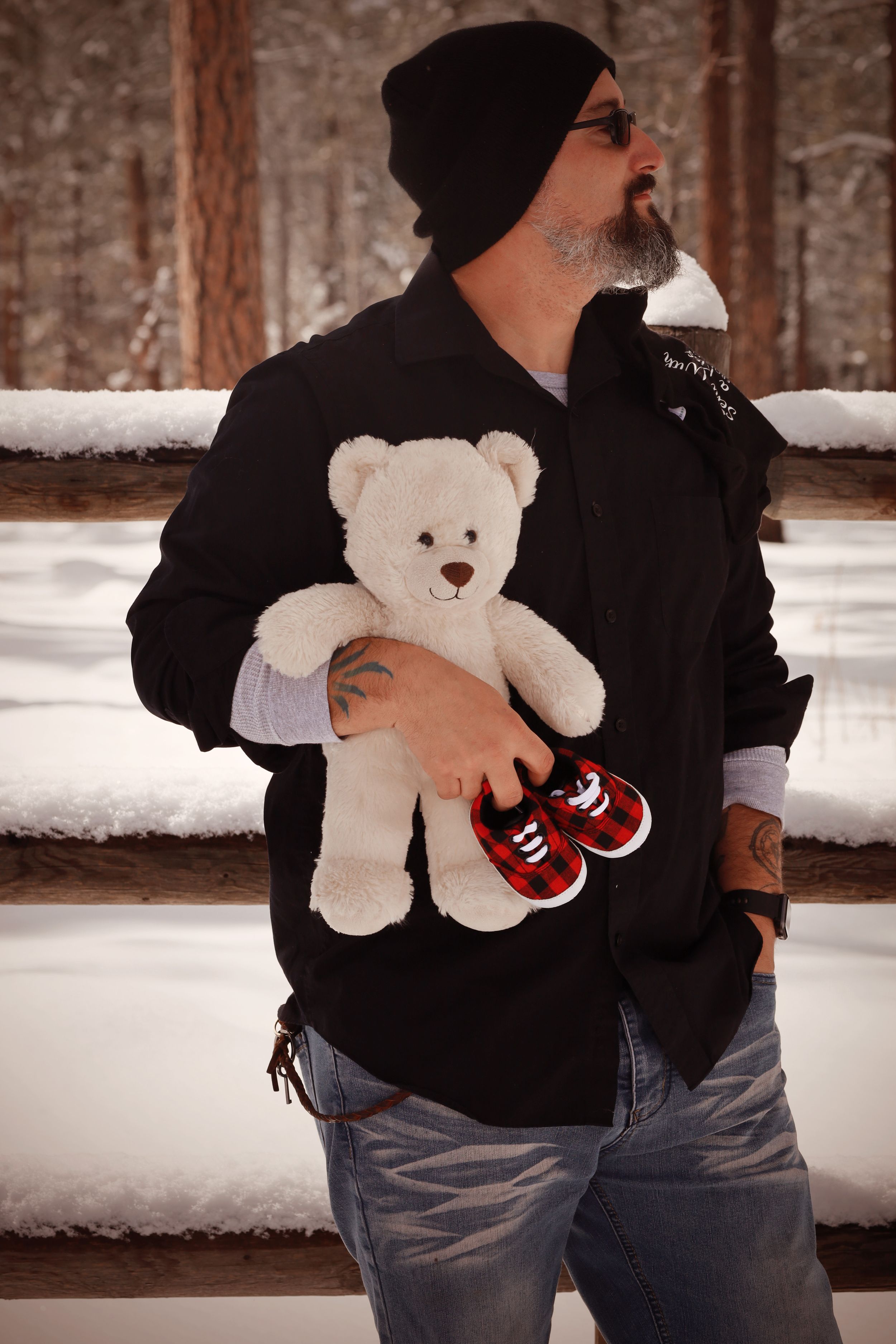Bearded man in black jacket and beanie holding white teddy bear with red baby sneakers in snowy setting.