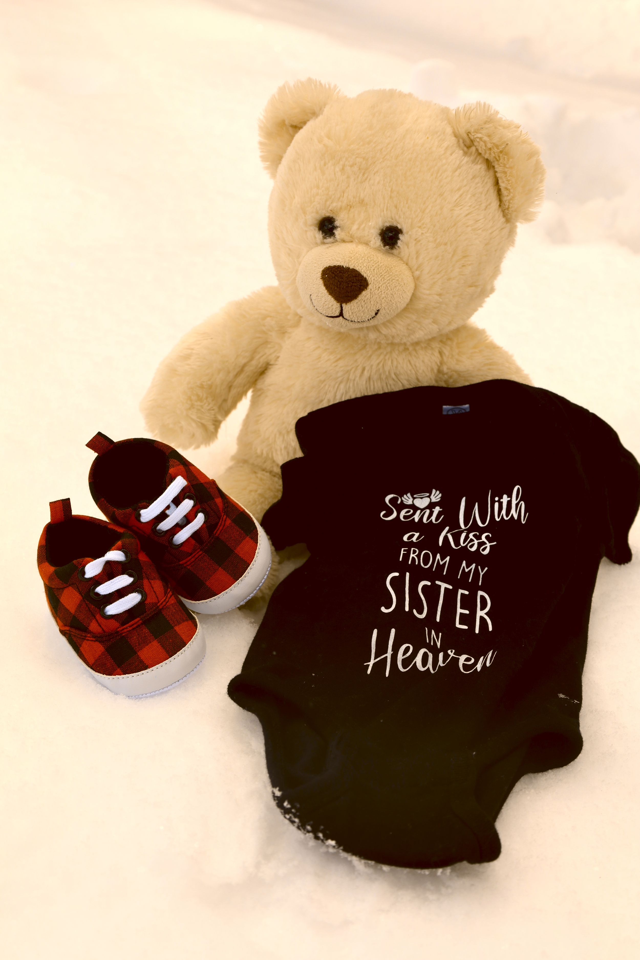 Black baby onesie reading Sent With a Kiss From My Sister in Heaven with plaid shoes and teddy bear in snow.
