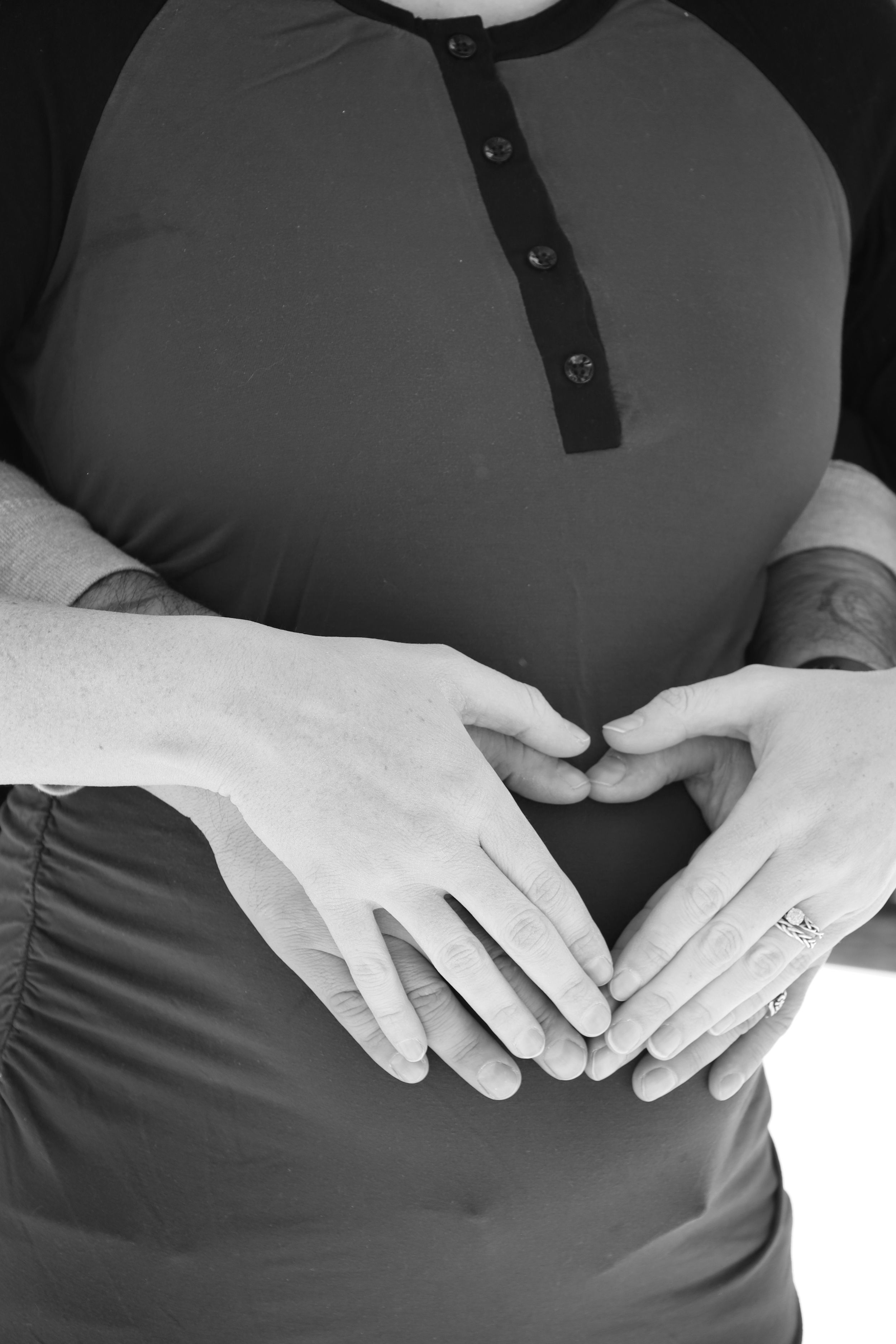 Black and white photo of pregnant couple forming heart shape with hands on baby bump.