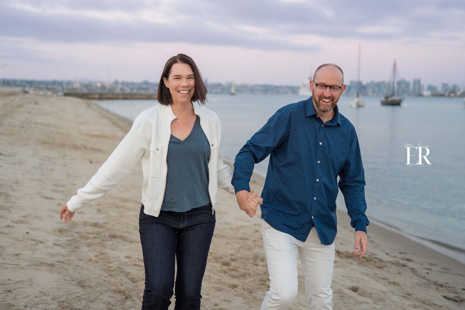 Couple walking along waterfront path with boats and city skyline in background.