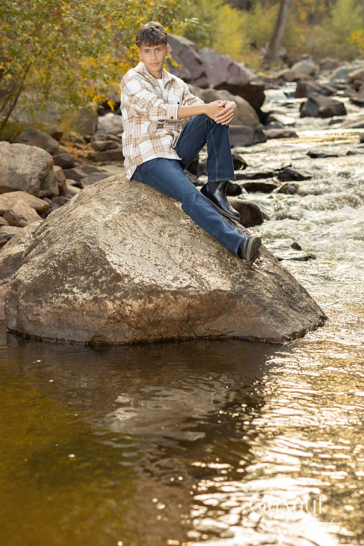Person in plaid shirt and jeans sits on large boulder by river during autumn with golden water reflections and rocks.