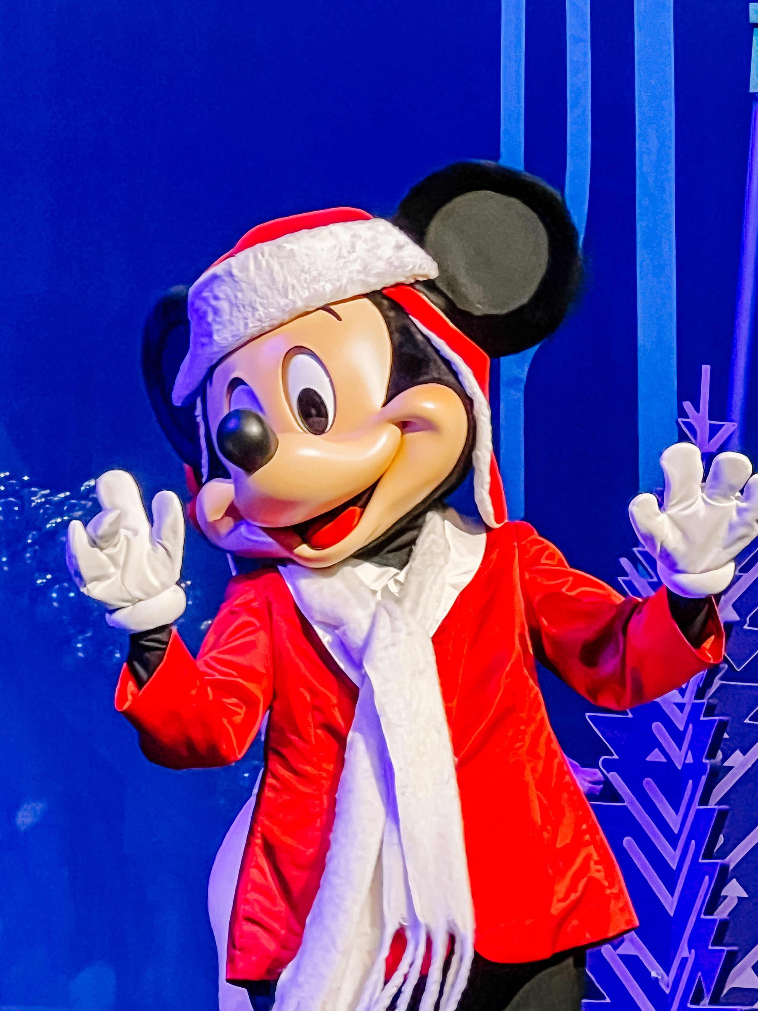 Mickey Mouse character in winter costume with red jacket and white gloves against blue background.