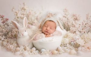 A sleeping newborn in a white bowl surrounded by delicate pink blossoms and a decorative white bunny figurine.