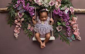 A baby sits under a floral arch made of pink and purple hanging flowers with greenery against a brown backdrop.
