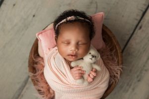 Sleeping newborn swaddled in pink wrap with toy teddy bear resting in brown wooden bowl against gray backdrop.