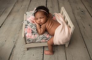 Sleeping newborn resting peacefully on floral cushion in rustic wooden bed with soft pink blanket.