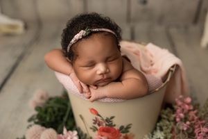 Sleeping newborn wrapped in pink blanket rests in decorative floral pot surrounded by lush greenery and pink flowers.