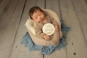 A sleeping newborn swaddled in beige fabric holds a round sign while lying on blue tulle fabric on wooden floor.